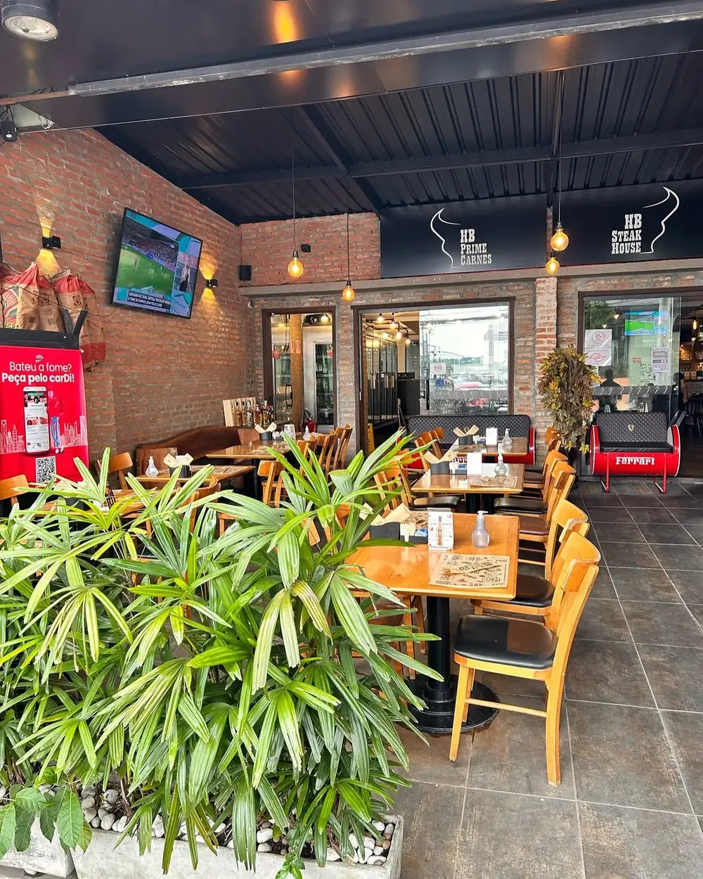 Salão da HB SteakHouse com mesas de madeira, cadeiras amarelas e tijolo aparente